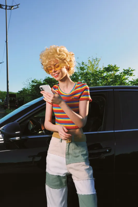 A stylish woman in a striped rainbow tee and patchwork denim stands beside a dark car, looking at a phone with a smile. Bright outdoor light highlights casual retro fashion.