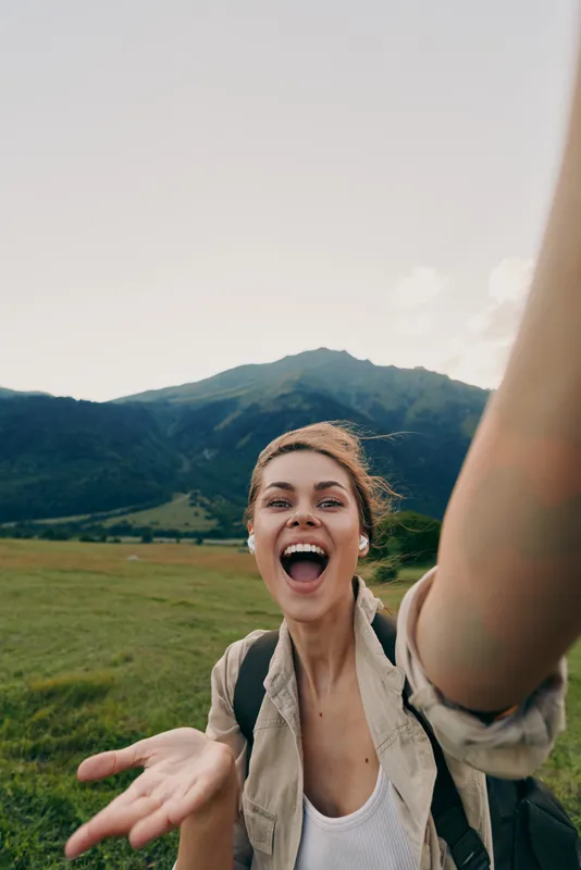 Young woman with backpack laughs into the camera in a sunny field with mountains in the background, capturing a carefree outdoor moment