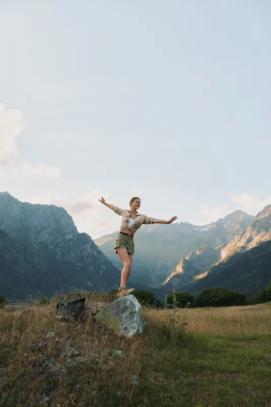 Cheerful woman balances on a rock in a sunlit mountain meadow, a carefree lifestyle moment amid wide landscape, clear sky, lush grass, distant peaks, and a gentle breeze.