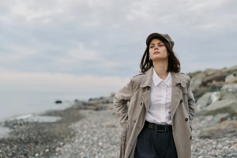 Young woman in trendy coat and hat stands on rocky beach with cloudy sky. She looks confident and thoughtful, with hands in pockets and natural makeup. Outdoor autumn fashion concept.