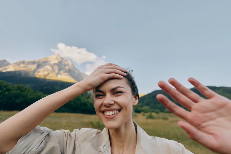 A smiling woman outdoors shields her eyes with her hand, sunlit landscape and distant mountains behind. A vibrant lifestyle portrait with natural light and carefree energy.