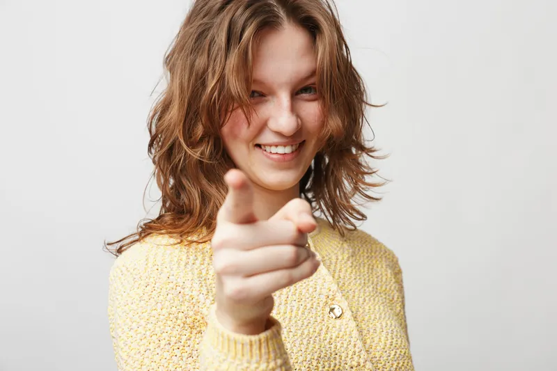 Smiling young woman with curly hair wearing yellow sweater points finger at camera. Friendly confident gesture on light background. Concept of communication and positive interaction.