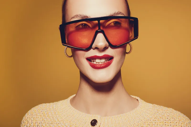 Smiling woman with red lipstick wearing large orange sunglasses and yellow sweater on yellow background. Fashion portrait with bright colors and confident expression. Concept of style and beauty.