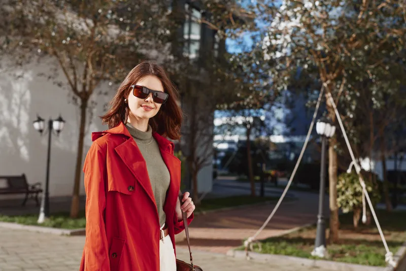 A stylish woman in a red coat walks through a sunny urban plaza, sunglasses on, exuding confidence and fashion in a casual street scene.