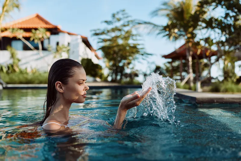 Young woman enjoys relaxing swim in outdoor pool splashing water with hand surrounded by tropical resort with palm trees and sunny weather lifestyle concept