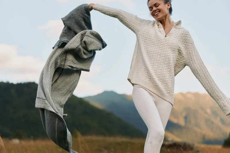 A joyful woman outdoors, twirling a coat as wind lifts her sweater, capturing movement in a sunlit countryside landscape, emphasizing freedom, nature, and casual fashion.