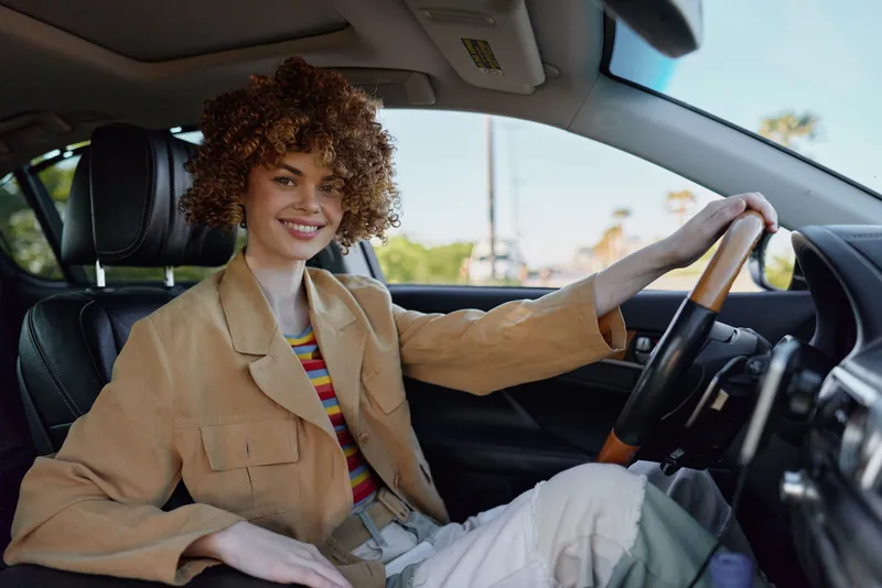 Smiling woman with curly hair sits in a car wearing a beige blazer and striped top. Bright natural light highlights casual chic style, urban lifestyle, and everyday travel moments with a retro film