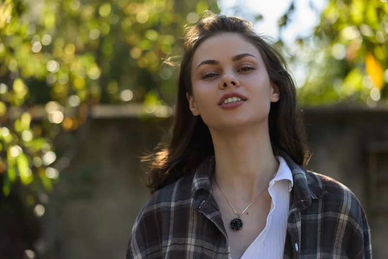 Woman in casual plaid shirt outdoors, natural light, relaxed smile, contemporary lifestyle portrait, greenery in background, simple necklace, candid mood, warm natural vibe, everyday fashion