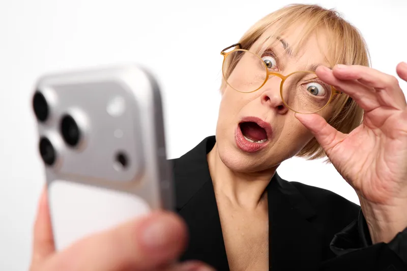 Surprised woman with glasses reacts to a shocked phone selfie, eyes wide, gaze intense, mouth open. Close up in studio lighting for technology amazement and excitement.