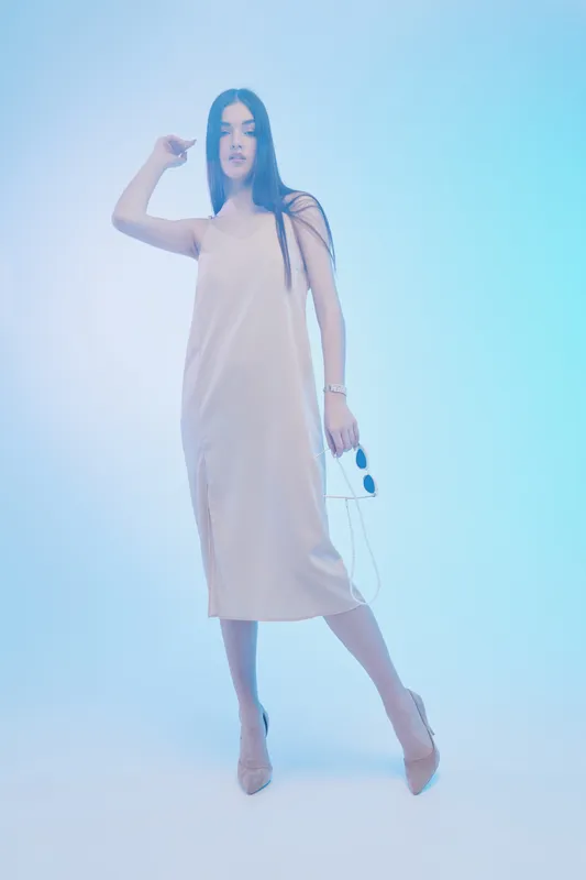 Young woman in a beige dress poses confidently against a soft blue background. Concept of beauty, fashion, elegance, and modern style in studio lighting.