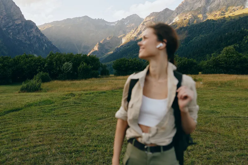 Young woman with short hair enjoys a mountain hike wearing casual clothes and earbuds while standing in a green meadow with forest and peaks in the background.