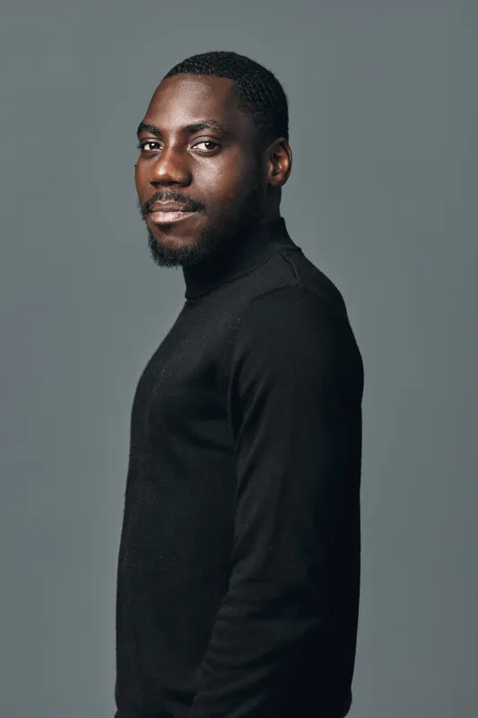 African man portrait in creative conceptual visual style wearing black turtleneck sweater on gray background. Confident young male with beard and short hair looking at camera.