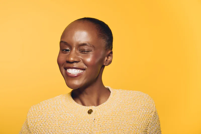 Smiling black woman with dark skin wearing a yellow sweater poses against a bright yellow background. The joyful expression and vibrant colors create a warm and positive mood.