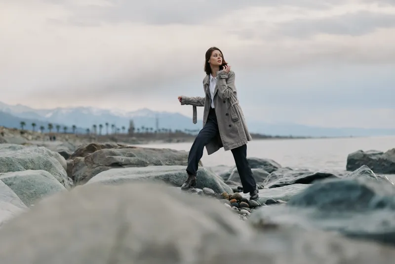 Young woman in checkered coat and black pants stands on rocks near the sea under cloudy sky. Casual fashion and nature concept with a calm coastal landscape in the background.