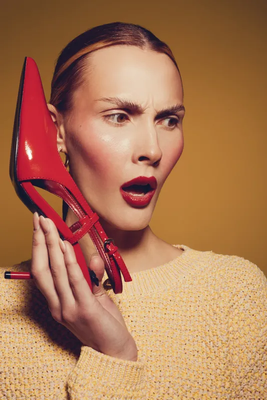 Surprised woman in yellow sweater holds red high heel shoe near face, looking shocked. Studio portrait with warm lighting on beige background. Concept for fashion, style, and emotions.