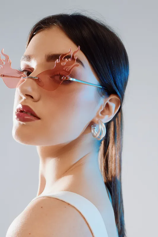 Young woman with sleek hair wearing stylish flame-shaped sunglasses and hoop earrings. Close-up portrait with modern fashion and beauty concept in studio lighting.