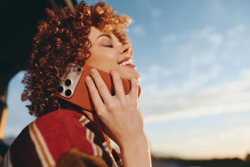 Woman with curly hair holds a smartphone to her cheek outdoors at sunset, smiling softly in warm golden light, casual lifestyle portrait, shallow depth of field, sky background