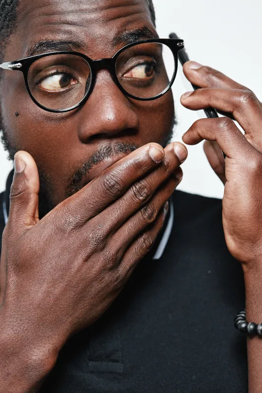 African American man with dark skin wearing glasses and black shirt covering mouth with hand in surprise or shock expression on white background.