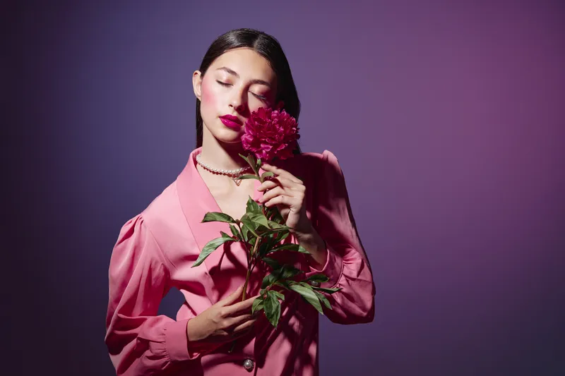 Elegant woman in a pink jacket holds purple flowers close to her face with closed eyes. Soft studio lighting and stylized makeup highlight beauty and creative fashion concept.