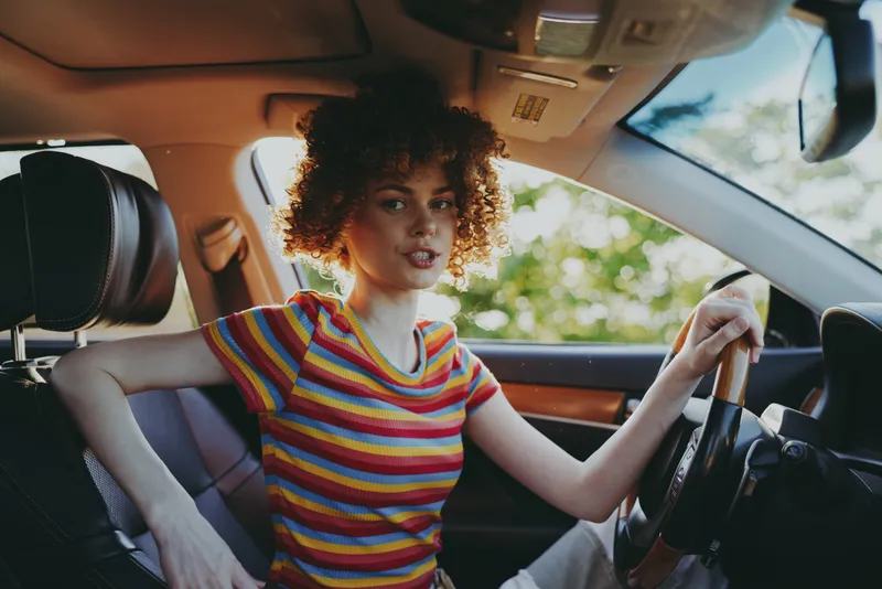 Cheerful woman in a colorful striped shirt sits in a sunlit car, relaxed posture behind the wheel, retro film color tones, natural light highlights her smile, casual lifestyle moment, breeze.
