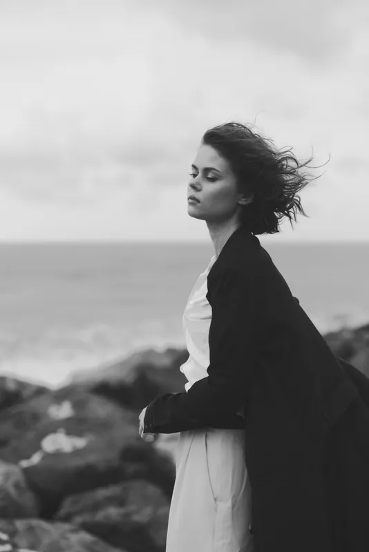 Woman standing along the sea in a flowing dress and dark coat, wind sweeping her hair, monochrome mood, coastal landscape, serene and contemplative, minimalist elegance and quiet strength