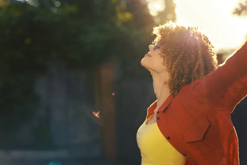 Young woman with curly hair raises her arms in a sunlit outdoor moment, wearing a yellow top and orange jacket, exuding freedom and playful confidence