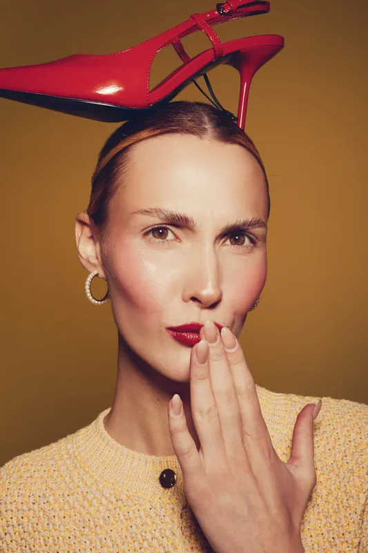 Woman with red high heel shoe on head poses with hand near lips, wearing yellow textured top and hoop earrings. Studio portrait with warm background, fashion and playful concept.