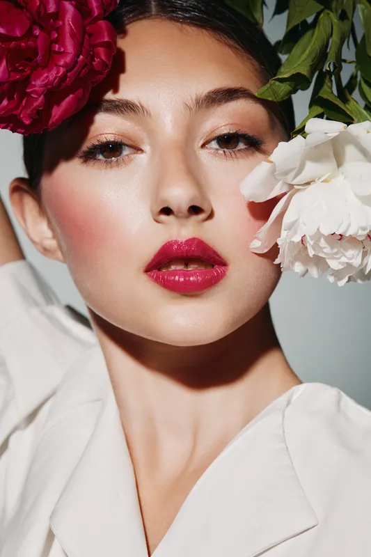 Close up portrait of fashionable woman with bold red lipstick and flowers by her face. Vibrant beauty, creative makeup, and clean white outfit in natural light for fashion theme.