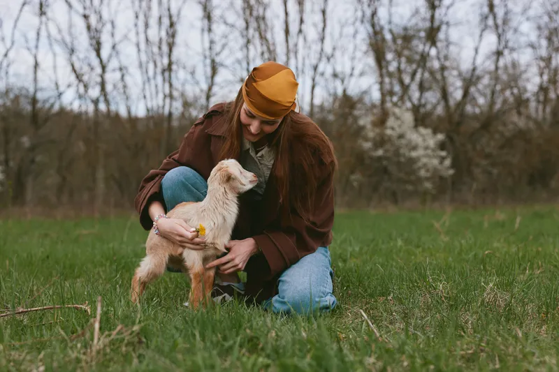Smiling woman kneeling in a green field, gently holding a baby goat. Natural outdoor setting with soft sunlight. Lifestyle moment emphasizing animal companionship, care, and wellness. Warm, intimate