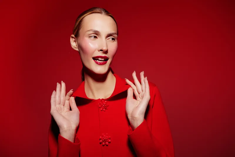 Young woman in red sweater poses with hands raised against red background. She has slicked back hair and bright red lipstick. Concept of fashion, style, and confident female portrait.