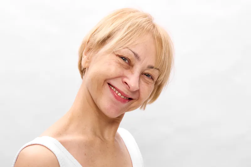 Woman smile portrait elderly blonde headshot friendly close up indoors on white background, soft light, short hair, relaxed posture, gentle expression, minimal composition