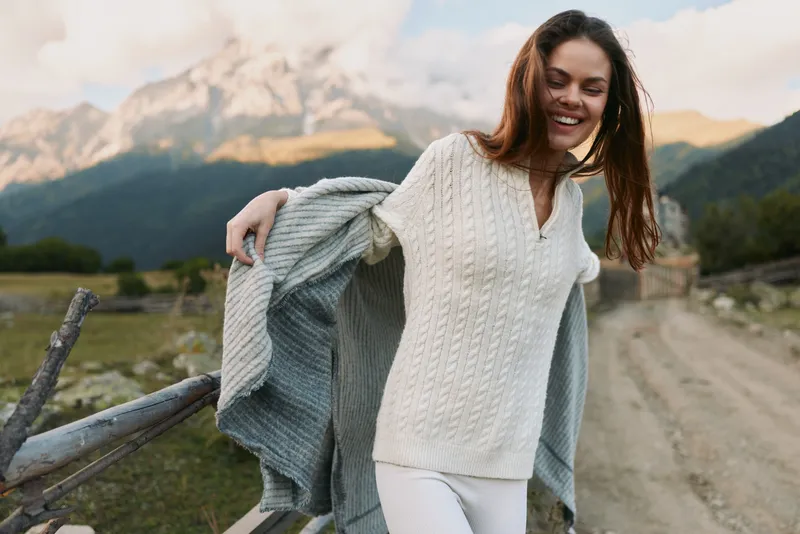 Woman enjoying outdoor lifestyle on a scenic mountain path, smiling, wearing a knitted sweater with a light jacket draped over her shoulders, open countryside and blue sky.