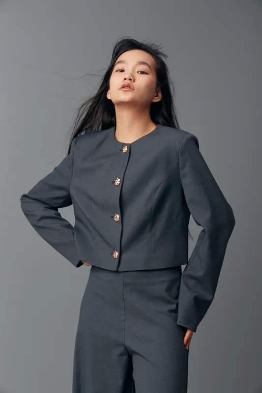 Young woman posing confidently in dark gray business suit with decorative buttons against neutral background. Fashion portrait of professional female model with long hair and serious expression.