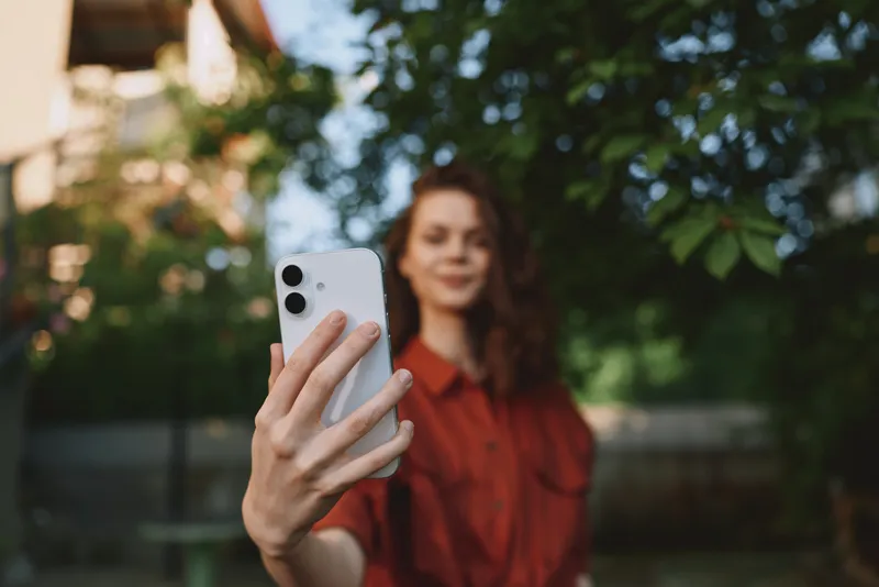 Smiling woman in a red blouse takes a selfie outdoors with a smartphone. Lush greenery and warm sunlight create a casual lifestyle moment focused on connection and everyday fun