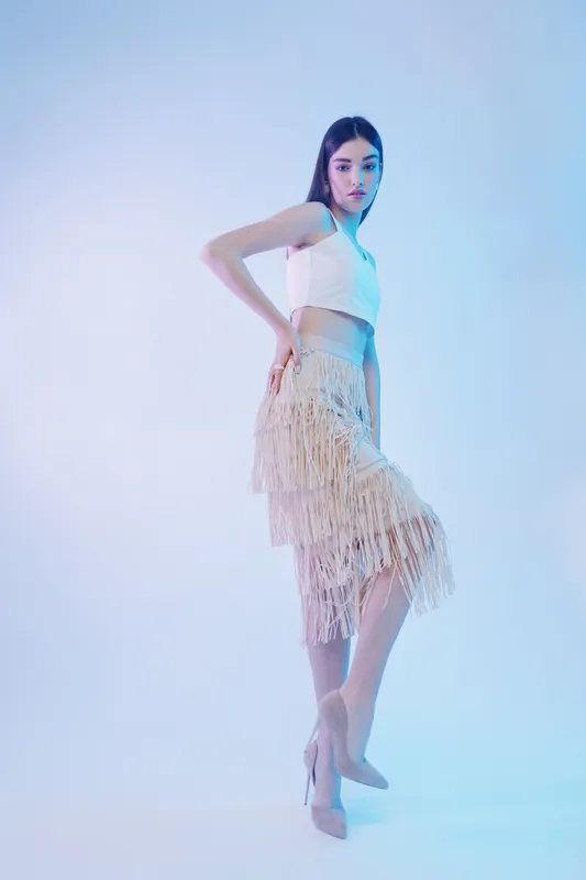 Young woman beauty fashion model poses confidently in a white crop top and fringed skirt on a soft gradient background. Concept for style, elegance, and modern trends.