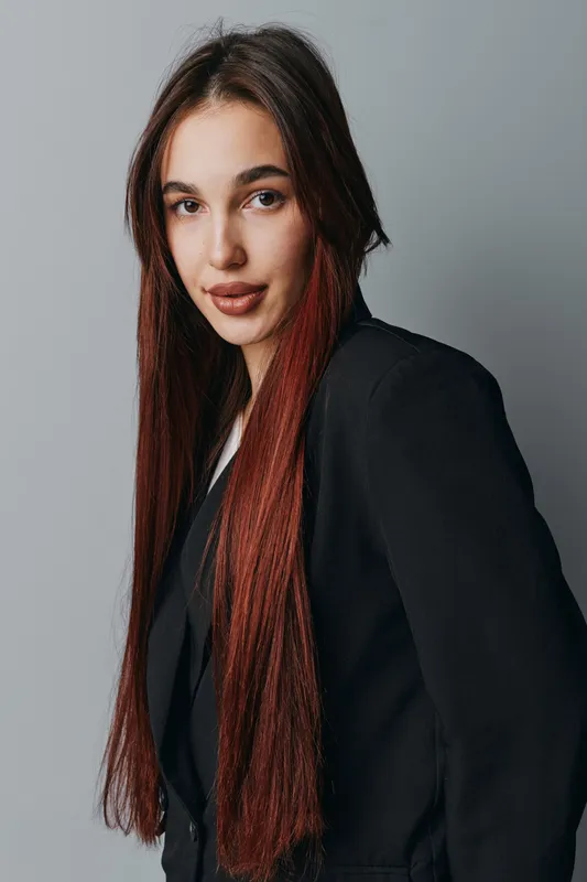 Young woman with long straight hair wearing black blazer posing confidently against gray background in studio. Female model with natural makeup and serious expression looking at camera.