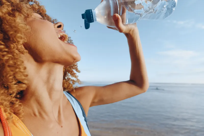 Woman with curly hair drinking water outdoors in summer by the ocean, hydration and wellness concept, close profile, sunny light, refreshment for healthy lifestyle