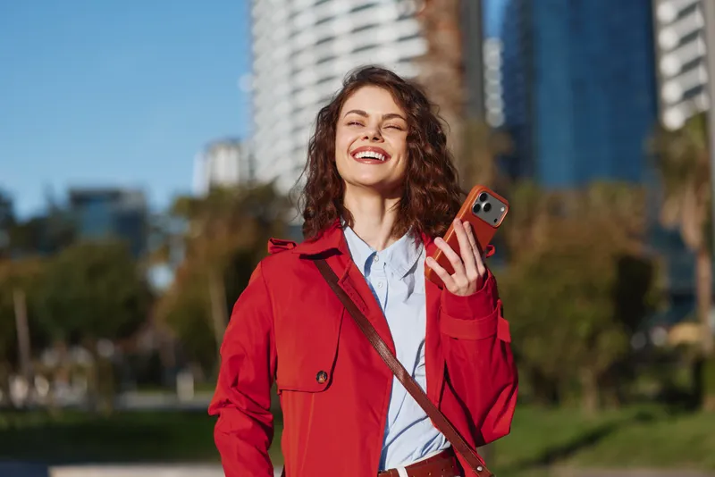 Young woman in a red jacket laughs outdoors in a sunny urban park, holding a smartphone and enjoying a carefree moment with a light breeze and bright city backdrop.