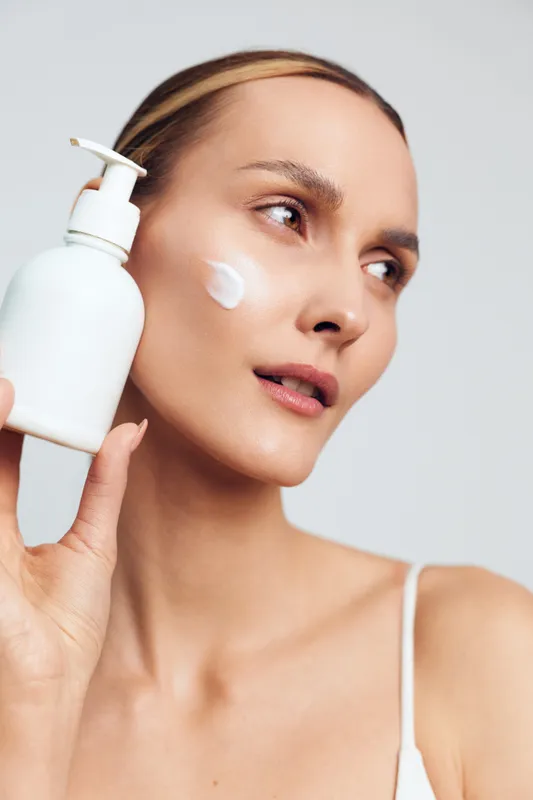 Young woman applies moisturizing cream on face holds white cosmetic bottle with dispenser has clear skin natural beauty isolated on light background skincare concept.
