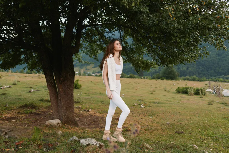 Young woman in a white outfit stands beside a large tree in a grassy countryside, looking upward with a serene, reflective expression in natural light.