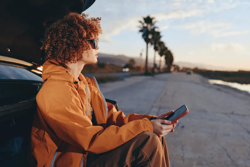 Woman lifestyle, outdoor travel scene at sunset, curly haired woman in orange coat sitting by car, holding smartphone and using map on road, calm smile, golden evening light
