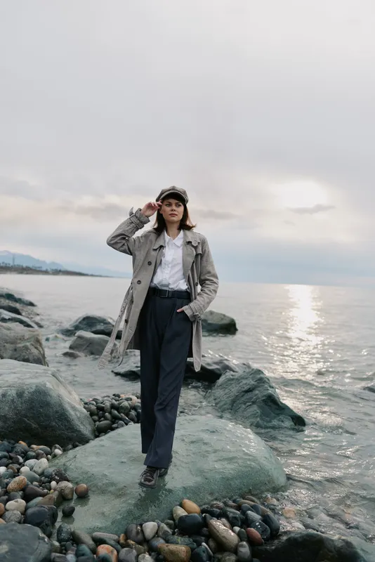 Woman in coat and hat stands on rocky shore near water under cloudy sky. Casual fashion, autumn style, and outdoor mood with calm sea and natural landscape background.