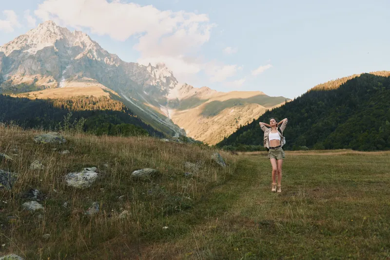 Young woman jumping with joy in a mountain meadow under blue sky. Concept of lifestyle, freedom, nature, and outdoor adventure. Bright natural light enhances the energetic mood.
