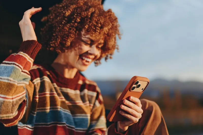Smiling woman with curly hair in a striped sweater holding a phone outdoors, taking a selfie at sunset, warm lifestyle moment, candid laughter, mountains softly blurred, natural light