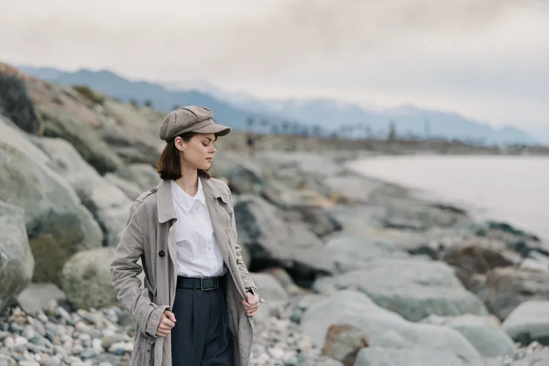 A young woman in a patterned coat and cap stands on rocky shore, looking thoughtfully at the sea. The cloudy sky and distant mountains create a calm, contemplative mood.