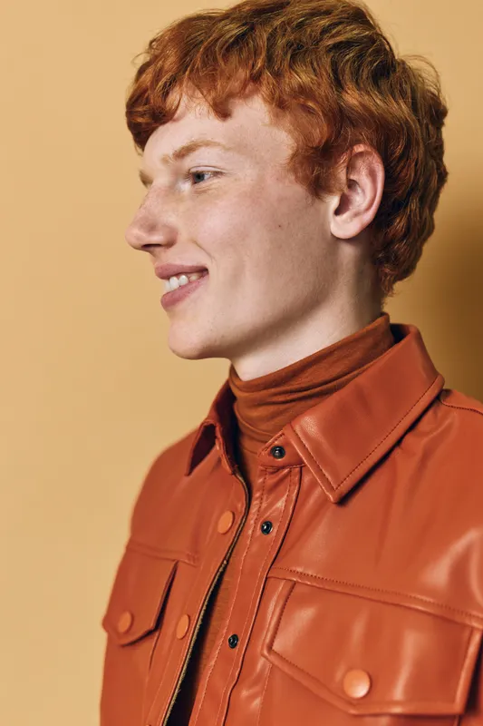 Young man with red hair wearing brown leather jacket and turtleneck smiling looking sideways on beige background in studio portrait profile view.
