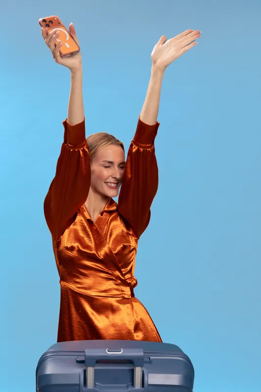 Smiling woman in orange satin dress raises hands holding smartphone with orange case above blue suitcase. Isolated on blue background. Concept of travel, joy, vacation, and lifestyle.