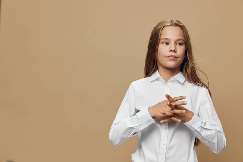 Girl with long hair wearing white shirt standing with clasped hands on beige background. Child looking aside with thoughtful expression for education or childhood concept.