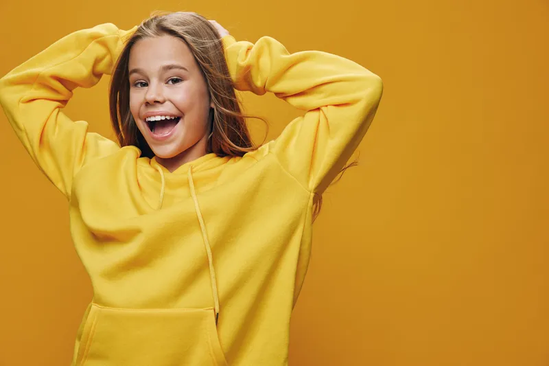 Happy girl in yellow hoodie smiling with raised hands behind head on vibrant orange background. Youthful cheerful female, casual style and positive mood for lifestyle and fashion.