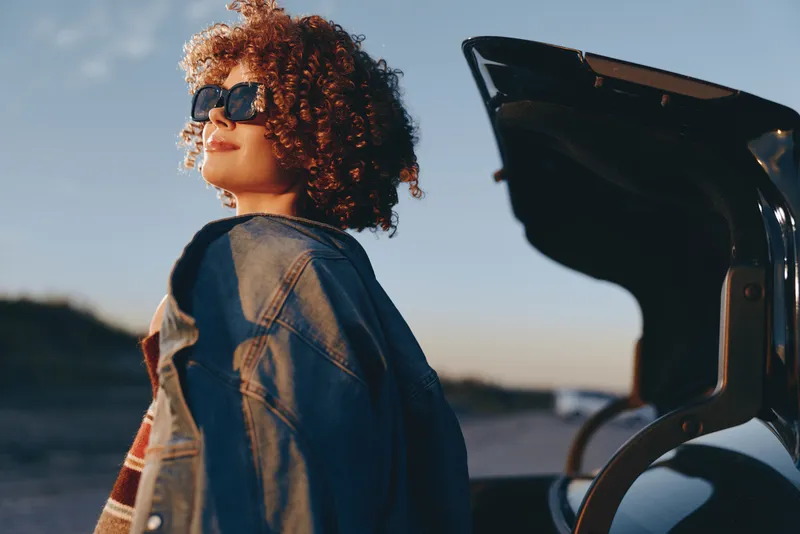 Woman lifestyle photo, curly hair, sunglasses, denim jacket, side profile near ocean, bright sunlight, calm travel outdoors on coastal beach path, gentle breeze, relaxed expression, focus